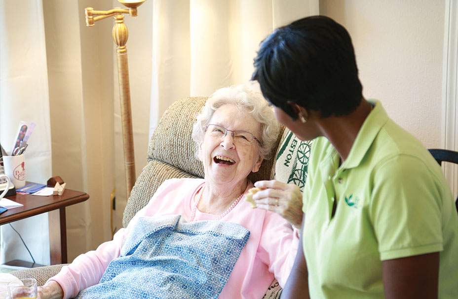 Caregiver leans over, speaking to her smiling senior client