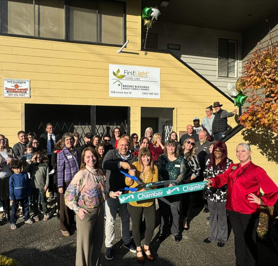 a group of smiling individuals stand for a ribbon cutting. The business building is behind. New Owners hold oversized scissors as they cut a ceremonial ribbon.