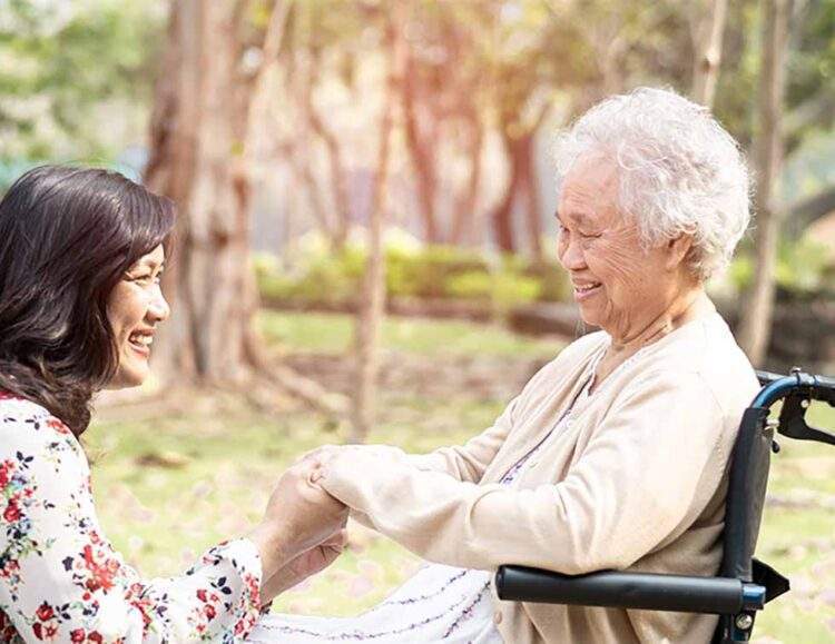 An adult daughter holds the hand of her mother, who is in a wheelchair